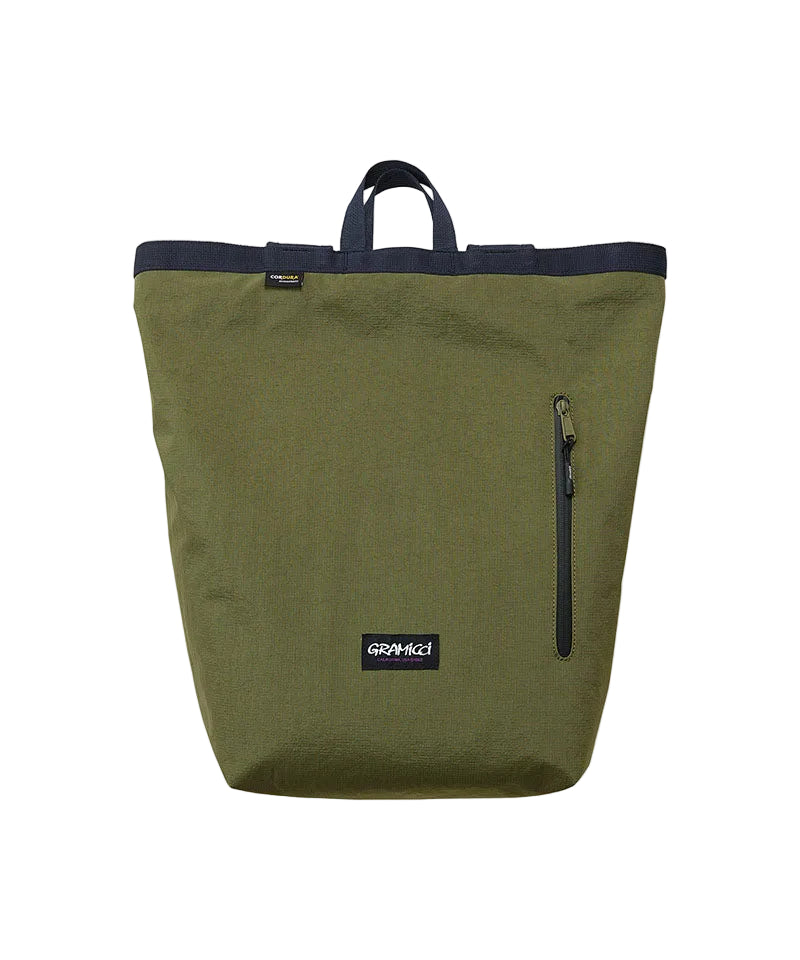 An olive green canvas tote bag with a dark navy blue top trim and handles, featuring a vertical zippered pocket on the side and a black brand patch.