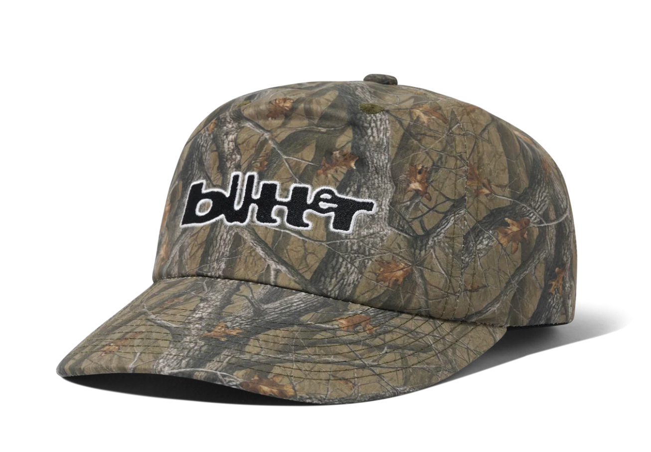 A camouflage baseball cap featuring a realistic woodland tree-bark and leaf pattern in shades of brown, green, and gray, with a black and white stylized 'blutt' logo embroidered on the front.