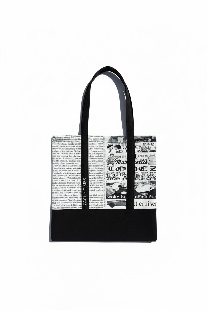 A black tote bag with a black-and-white newspaper print panel on the front, laid flat on a textured gray asphalt surface.